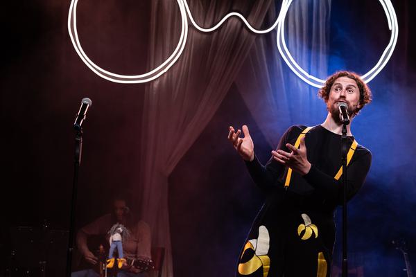 Gary McNair in Dear Billy stands on stage in front of a large pair of neon glasses.