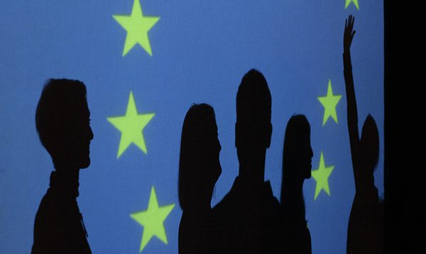 Five people are silhouetted against the European Union flag. Four stare straight ahead into the distance, whilst one person jumps up to reach for one of the stars on the flag.