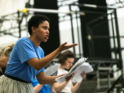Danielle is reading from a script with one hand stretched in front of her 	                gesturing with a look of anger. Catriona and Anne are in the   	b		   background but are out of focus.
