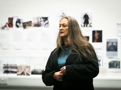 Liz looks to the side with both hands clasped in front of her. In the 			   background is blurred set and costume images on the rehearsal wall.