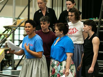 The cast stand around Danielle all looking at her, Danielle is holding a 		     script in her hand while reading an emotional piece of text.