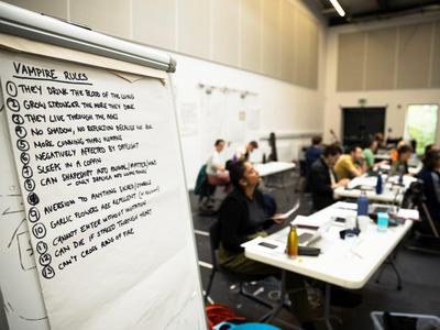 An out of focus shot of the Dracula creative and production team 		watching rehearsals, in focus is a flipchart with the 13 ‘rules’ of vampirism.