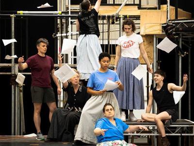 The cast stand and sit all over the set. Danielle is in the middle of the group. Ailsa has her back to everyone and is throwing sheets of paper into the air. There are papers strewn everywhere.