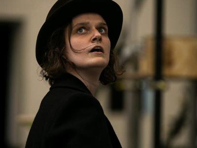 Catriona stands and looks upward in fear. She is wearing a black top hat and blazer.