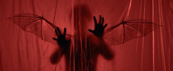 A shadowy figure with bat wings lurks behind a red translucent curtain