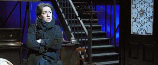 A pale woman with dark hair in a long black dress, coat and scarf stands at the end of the stage, looking out to the distance. To the right there is a door with a frosted glass panel letting light through, a coat stand, and a flight of stairs leading up to second level of the set.