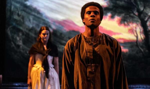 Two actors stand, partially cast in shadow, against a sunset backdrop