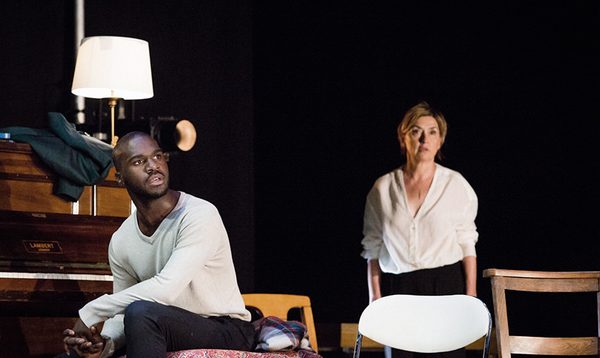 Thierry Mabonga and Isabelle Vincent in First Snow / Premiere Neige. Thierry sits on the left of the screen beside a piano, staring into the distance. Isabelle stands slightly out of focus behind him, also staring off into the distance.