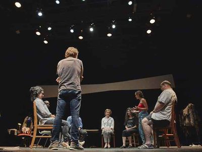 Most of the cast are sat facing each other on wooden chairs, two people are standing up.
