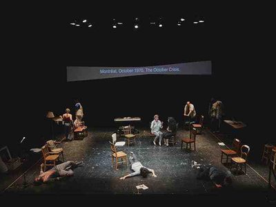 The cast are spread across the stage. Some sit on chairs. One is lying face up on a chair. Another two lie face down on the floor, as if they have collapsed.