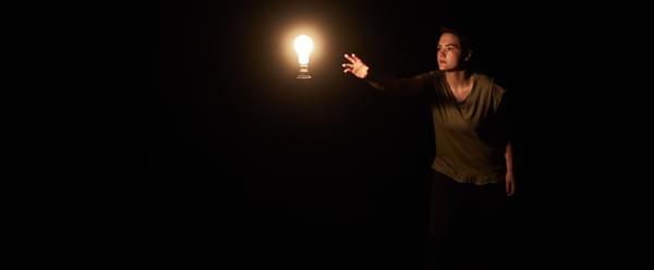 A person reaching towards a lit bulb in a dark room