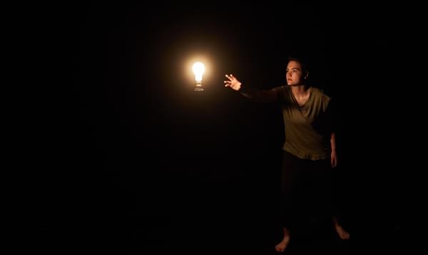 A person reaching towards a lit bulb in a dark room