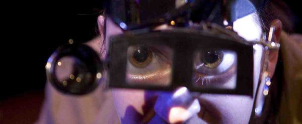 A woman wearing steampunk magnifying goggles looks into the camera. Her eyes are enlarged by the magnifying glass.