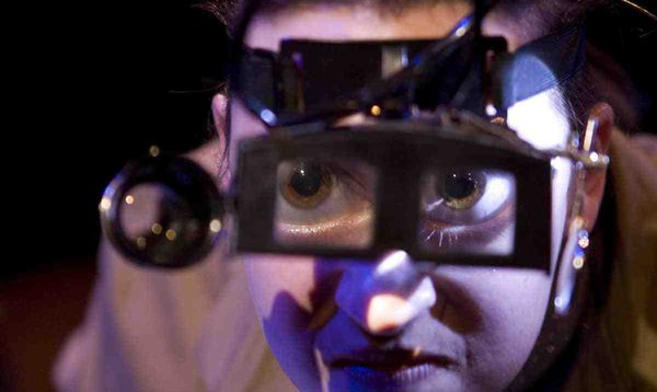 A woman wearing steampunk magnifying goggles looks into the camera. Her eyes are enlarged by the magnifying glass.