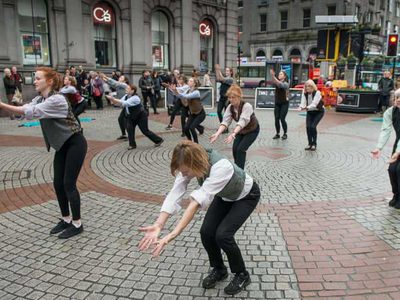 A group of dancers dance in formation in the middle of a busy shopping street