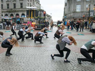 A group of dancers dance in formation in the middle of a busy shopping street