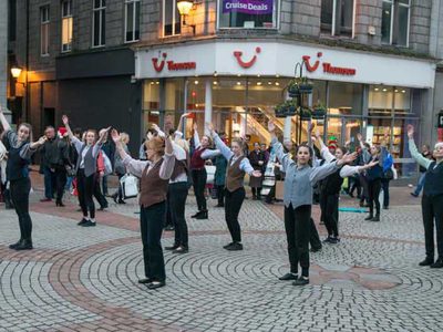 A group of dancers dance in formation in the middle of a busy shopping street