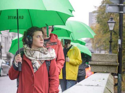A woman with a green umbrella walks down a street wearing headphones