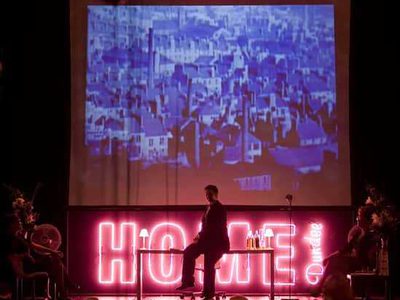 A pink neon sign spelling out HOME DUNDEE is illuminated against a black backdrop. Above this, a black and white photo of Dundee is projected above the sign. In front of this, an actor in a suit sits on a table.