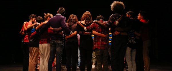 Actors stand on a stage with their arms around each others shoulders