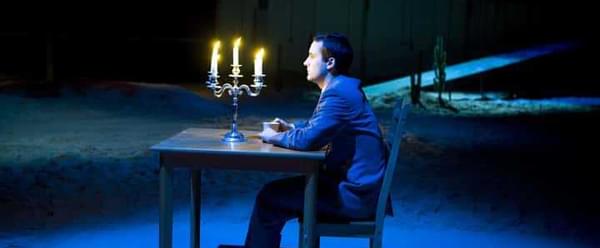An actor in a suit and trainers sits in a chair at a table. There is a candelabra with five lit candles on the table. The stage is covered in a blue light, casting a shadow behind them.