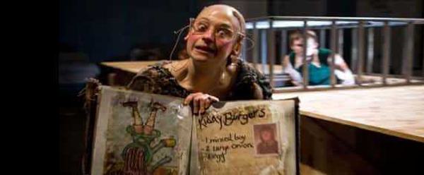 A balding woman wearing glasses holds a book towards the camera with a recipie for 'Kiddy Burgers'. The ingredients lists 'one minced boy', '2 large onions' and '1 egg'. Behind her, a boy is trapped in a cage.