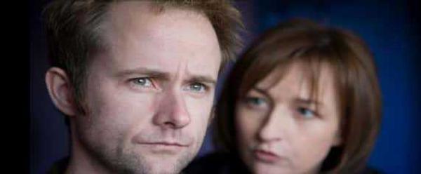 Two actors against a blue background. A man looks off into the distance, behind him a woman is looking at and talking to him.