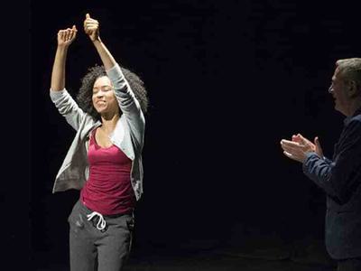 A young woman snaps her fingers in the air and dances. An older man looks on, clapping.