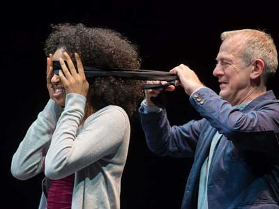 An older man puts a blindfold on a younger woman. They both smile.