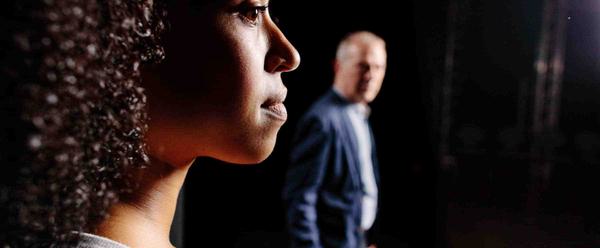 A young woman stands on a stage looking forward. Behind her, and slightly blurred, a man also standing on the stage looks towards her.