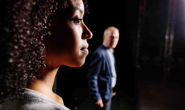 A young woman stands on a stage looking forward. Behind her, and slightly blurred, a man also standing on the stage looks towards her.