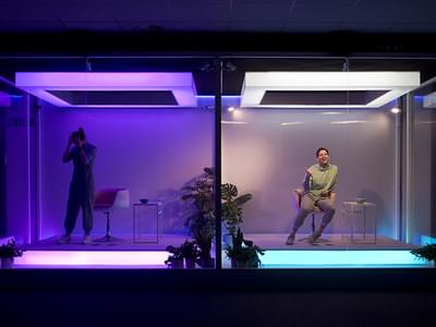 On the left a woman holds her head in her hands inside a purple-lit cube, on the right a man sits in a chair in a blue-lit cube