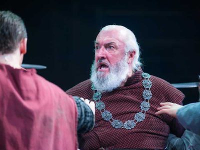 A man with white hair and a white beard is held at sword point. He looks angry.