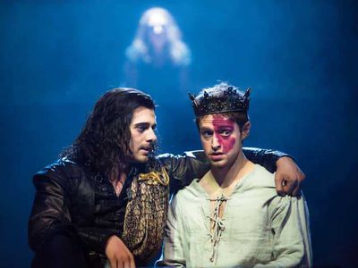 A man in battle-wear puts his arm around a young king with a birth mark on his face. The young king looks worried.