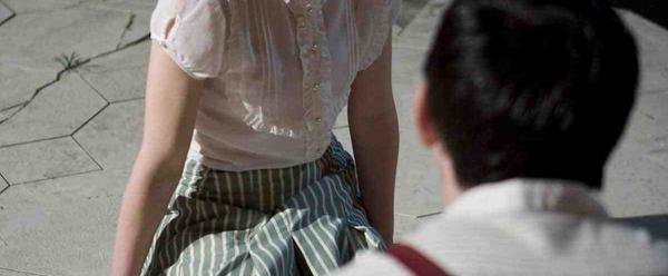 A young woman in a white shirt and striped skirt sits on the ground, facing the camera. She is looking towards a male actor in a white shirt who has his back to the camera.
