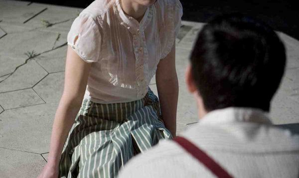 A young woman in a white shirt and striped skirt sits on the ground, facing the camera. She is looking towards a male actor in a white shirt who has his back to the camera.