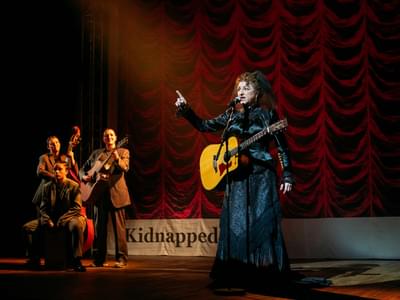 An actor wearing a mourning dress has a guitar and speaks into a microphone