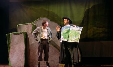 two men stand next to a boulder - man on right holds a map