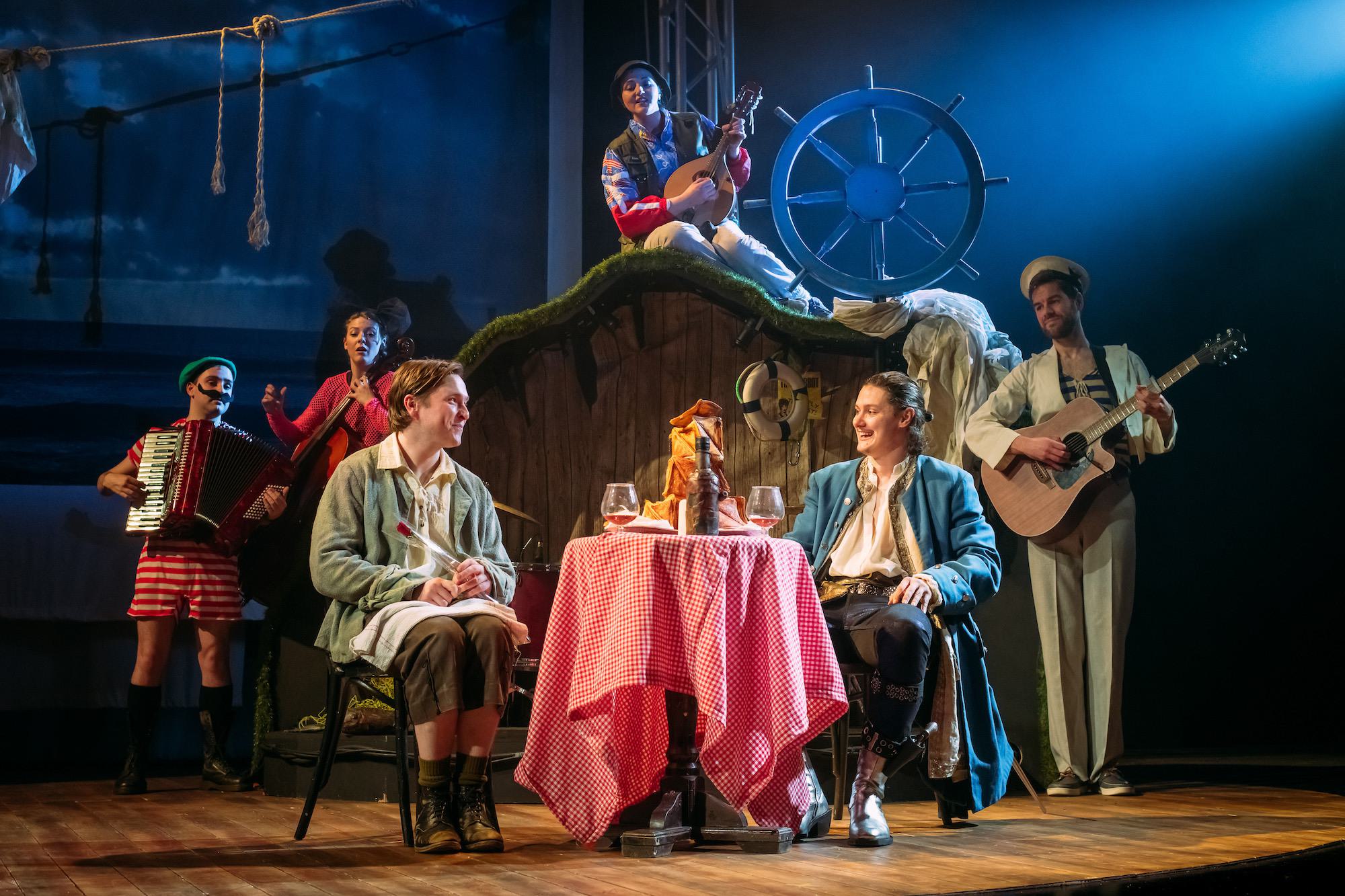 Photograph from the play, showing Alan and Davie sitting together at a dinner table smiling at each other; in the background are four sailors, who wear colourful anachronistic costumes and play musical instruments.