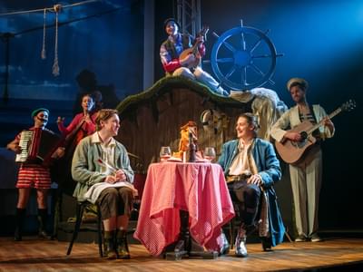 Two actors sit and drink at a table whilst an ensemble play instruments in the background