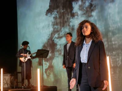 Two actors stand performing whilst a musician plays guitar in the background