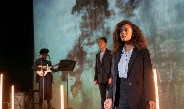 Two women stand on stage, with a third playing guitar in the background