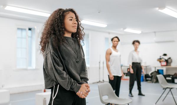 An actor in a grey sweatshirt stands with their hands clutched together