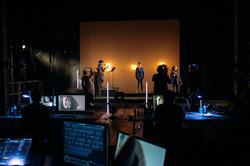 The foreground is a series of screens, at the back of the shot are the musicians and performers performing onstage