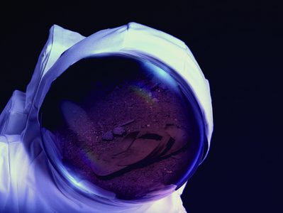 A spacesuit against a black background. Reflected in the glass of the astronaut's helmet there is a flipflop on a dusty terrain.