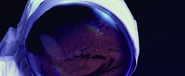 A spacesuit against a black background. Reflected in the glass of the astronaut's helmet there is a flipflop on a dusty terrain.