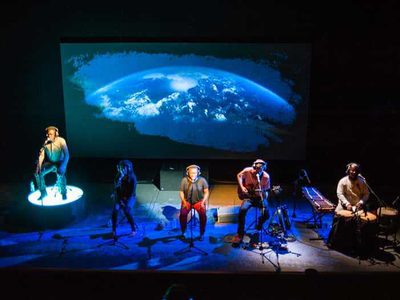 Men and women sit on chairs in a row in front of microphones. A blue lit moon can be seen behind them.