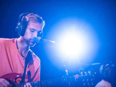 A man wearing headphones plays a guitar on a blue lit stage