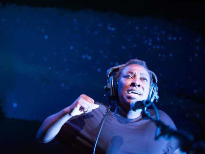 A woman wearing headphones speaks animatedly into a microphone on a deep blue stage