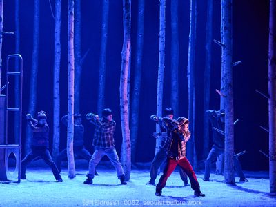 Six actors stand with there legs apart. Each one holds a knife and holds their arms high, as if to stab something. They almost look like they are dancing. The lighting is blue and the actors are woven amongst trees.